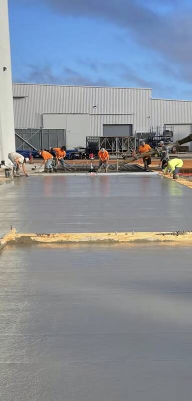Crew screeding and floating a large wet concrete slab in front of a metal warehouse, with a pump hose at the right