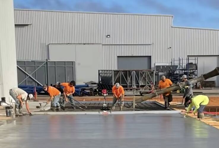 Crew in orange and hi-vis shirts finishing a large fresh concrete slab with a pump hose in front of a metal building