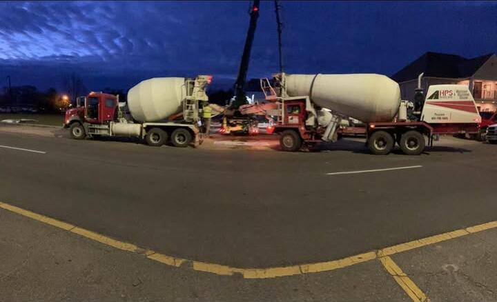 Two ready-mix trucks discharging concrete at dusk, a boom between them and a worker in a hi-vis vest