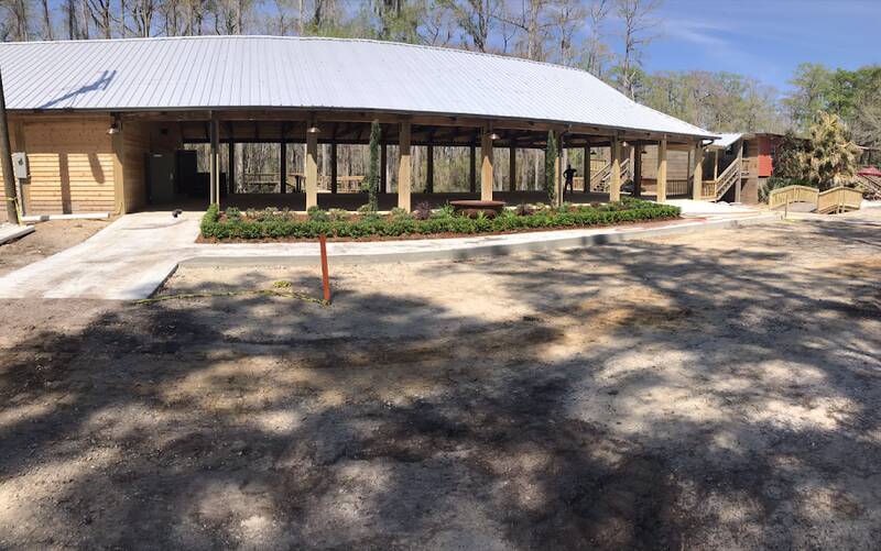 Open-sided pavilion with a metal roof and wood posts, a concrete walkway at left and a concrete curb along a planted bed